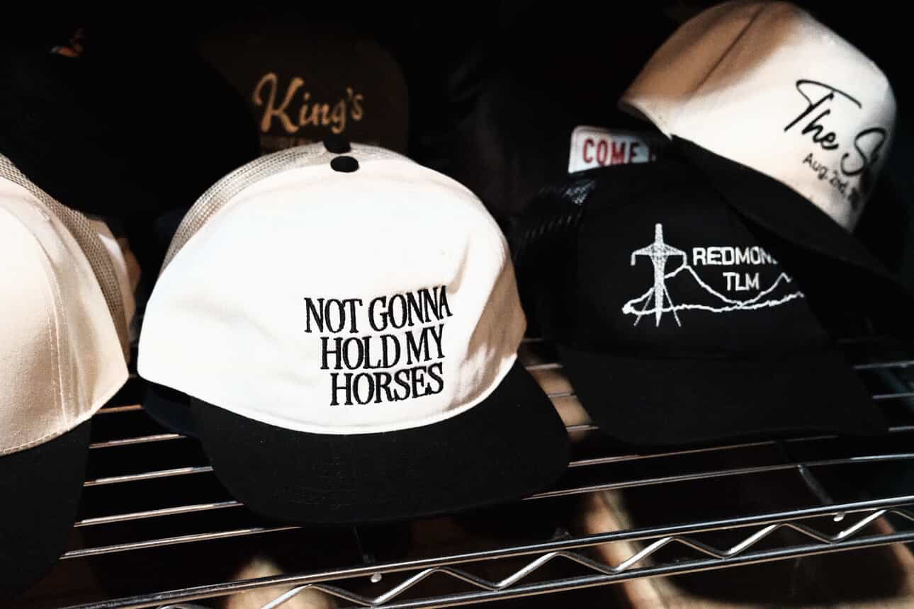 White and black baseball cap with text, displayed on a metal shelf, surrounded by other caps, some with logos and text.