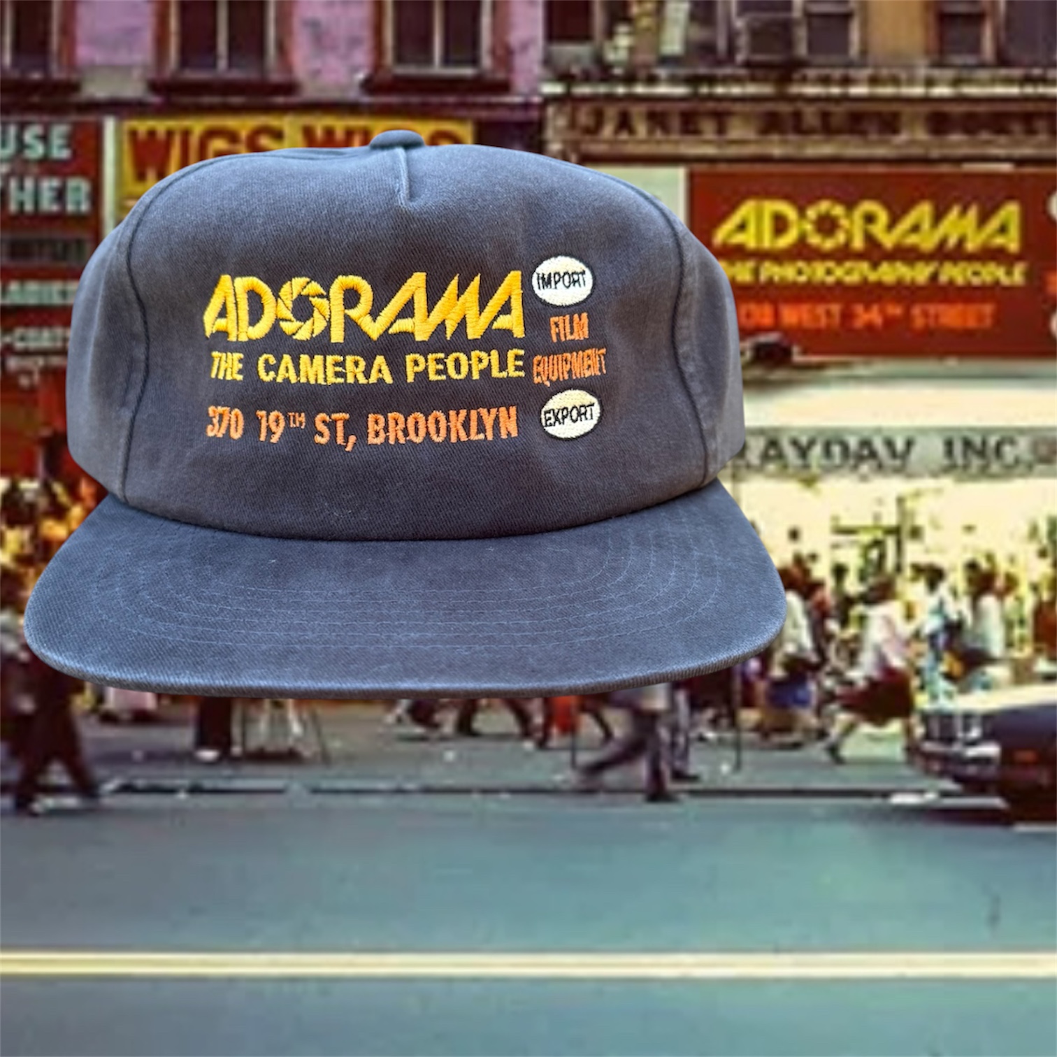 Gray baseball cap with yellow embroidered text, in front of a busy city street scene with storefronts and pedestrians.