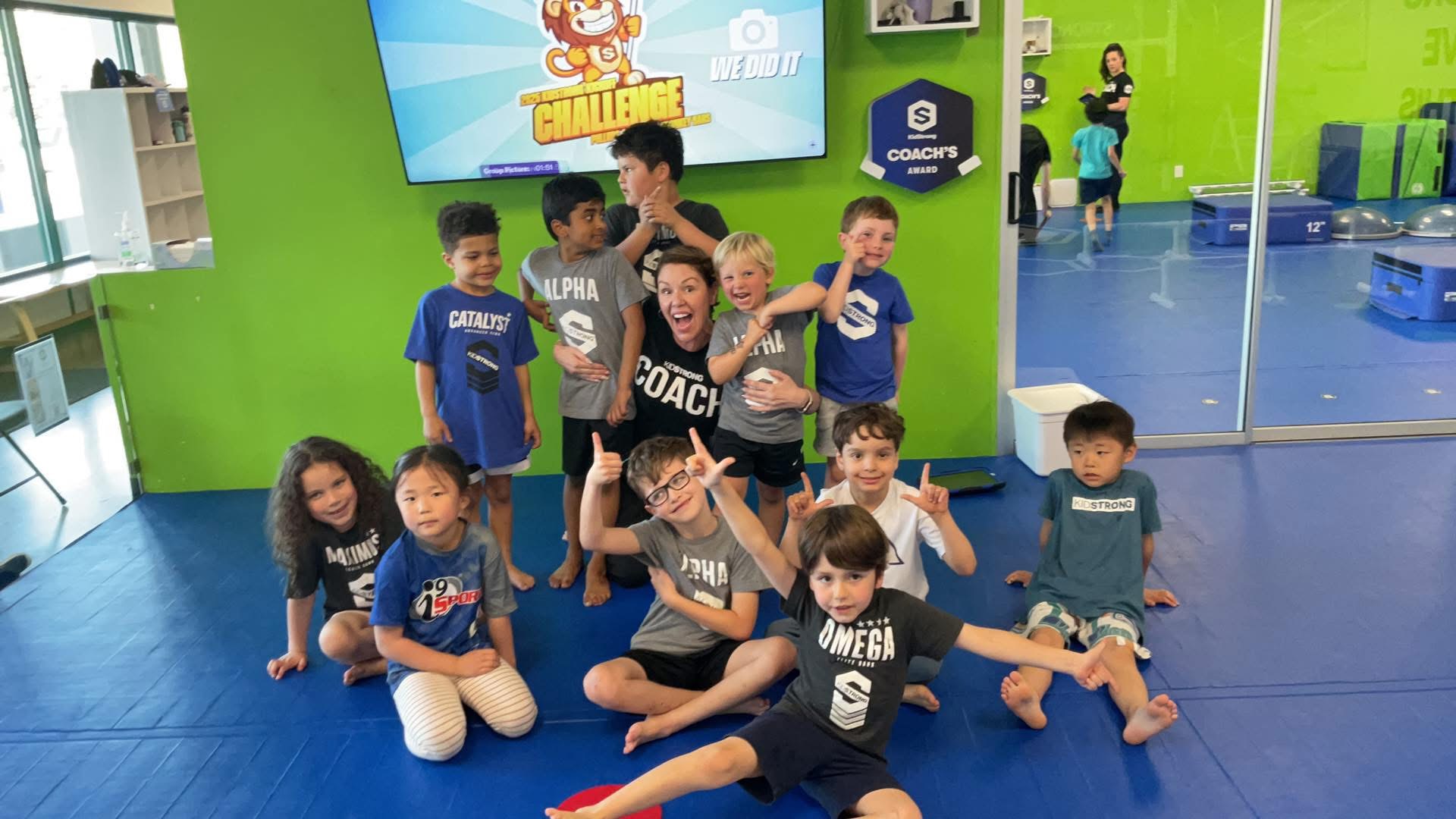 Group of children and coaches in a colorful indoor play area, some sitting and some standing, with a green wall and a screen.