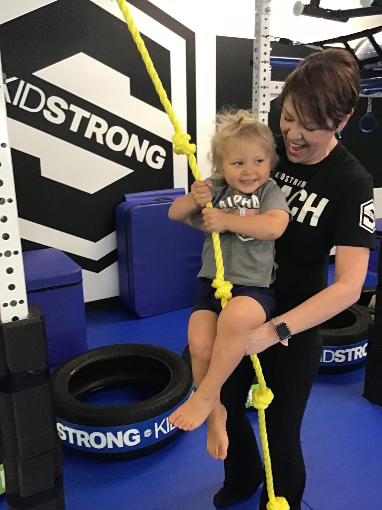 Child sitting on a trainer’s lap holding a yellow rope in a gym, with gym equipment and a sign in the background.