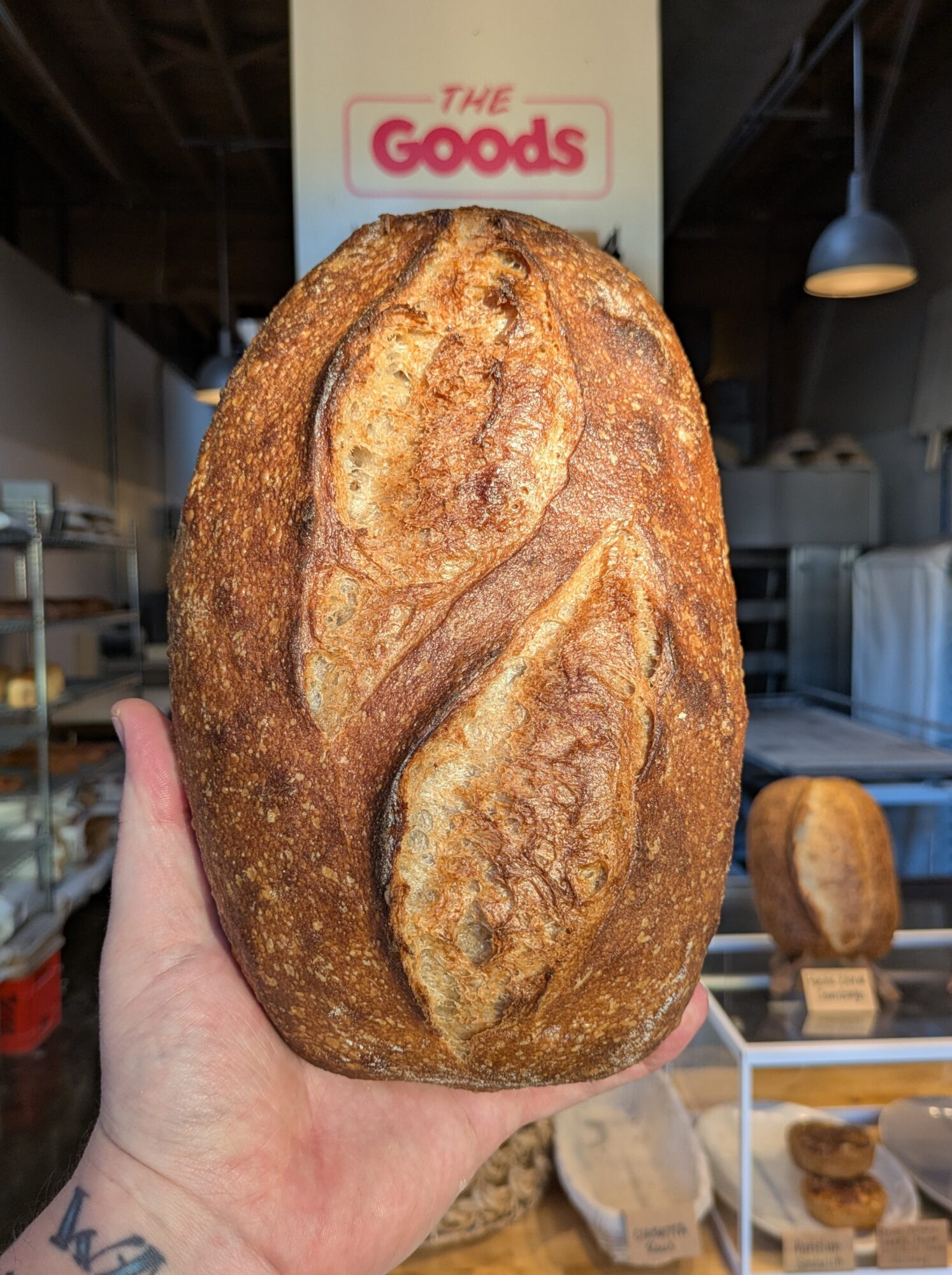 Hand holding a large, oval-shaped loaf of bread with a crusty surface and decorative scoring, in a bakery setting.