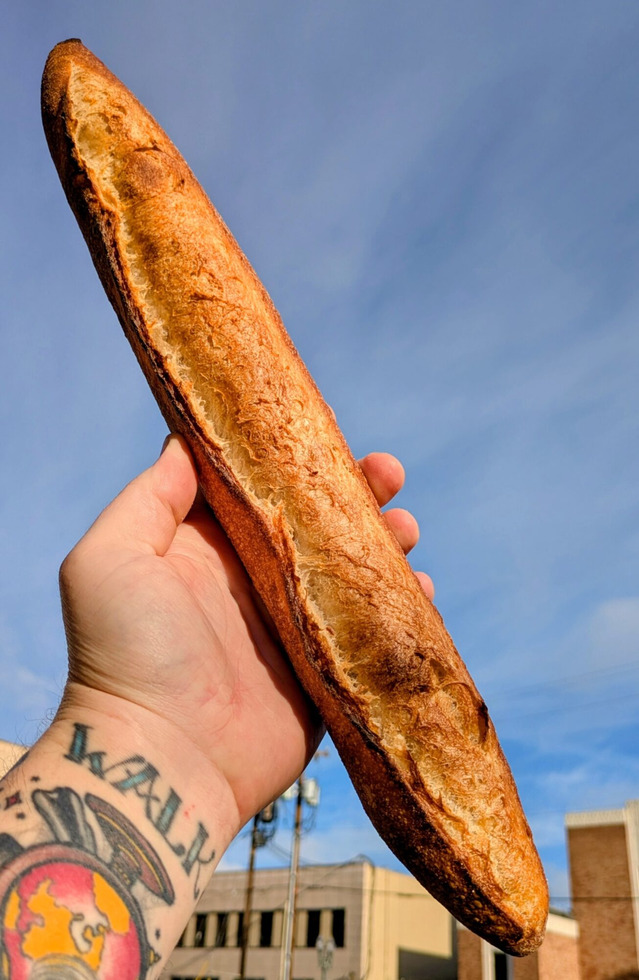 Person holding a long, toasted baguette against a blue sky background, with a tattoo visible on their wrist.