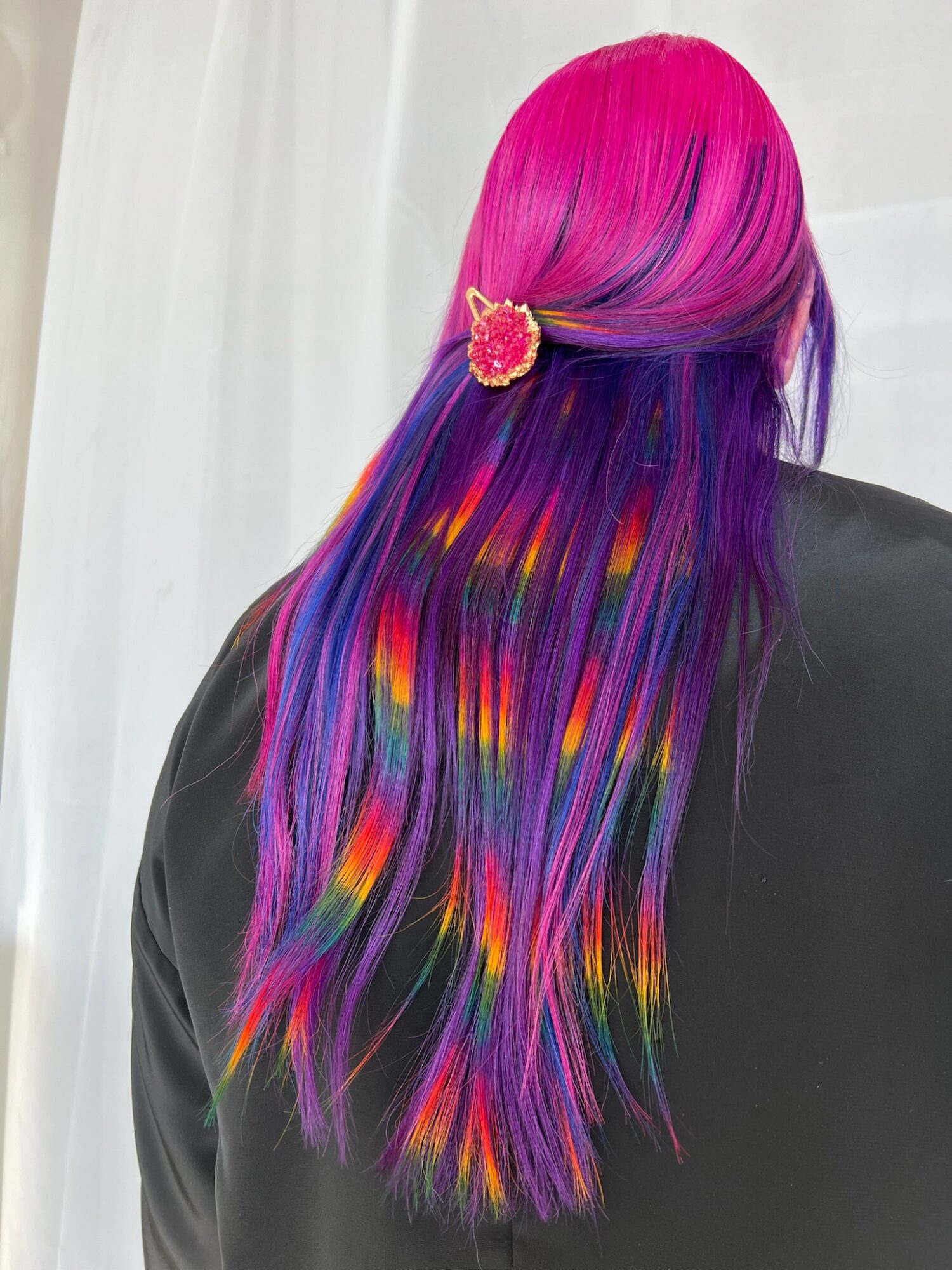 Back of a person with long, rainbow-colored hair and a pink hair clip, wearing a black jacket, against a white background.