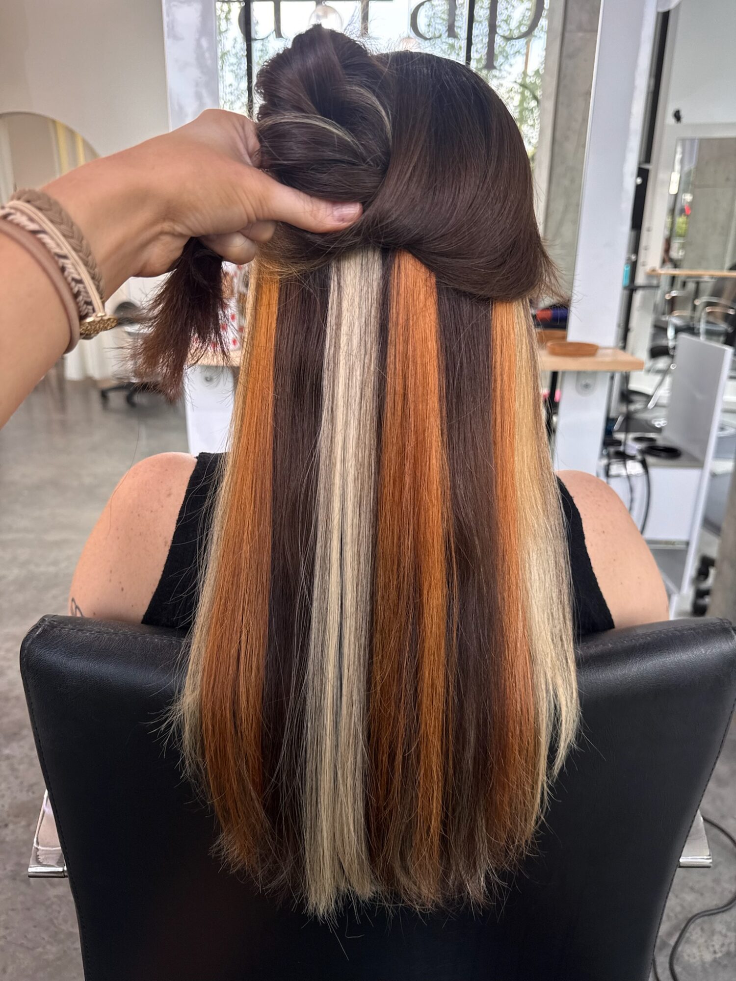 Back view of a woman with long, straight hair featuring multiple colored streaks, seated in a salon chair.