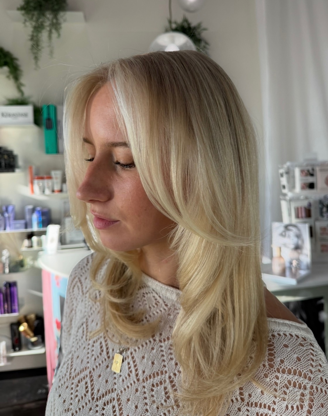 Woman with long blonde hair and a lace top, looking down in a well-lit room with shelves and products.