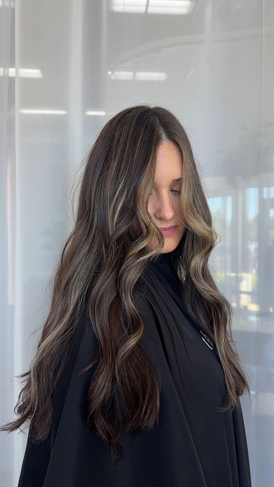Woman with long wavy hair looking down, wearing a dark top, standing indoors with a glass background.
