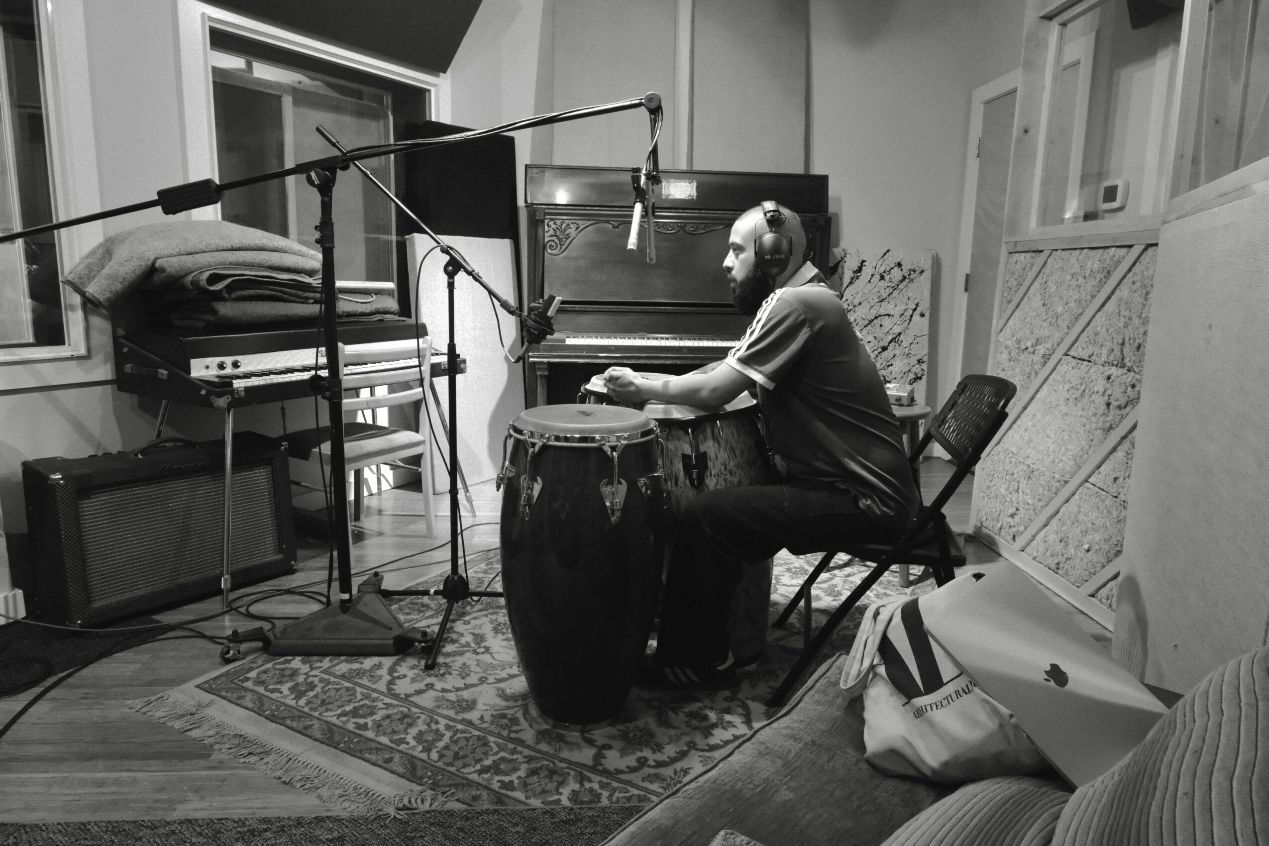 Person wearing headphones plays a drum in a recording studio with microphones and equipment, sitting on a chair.