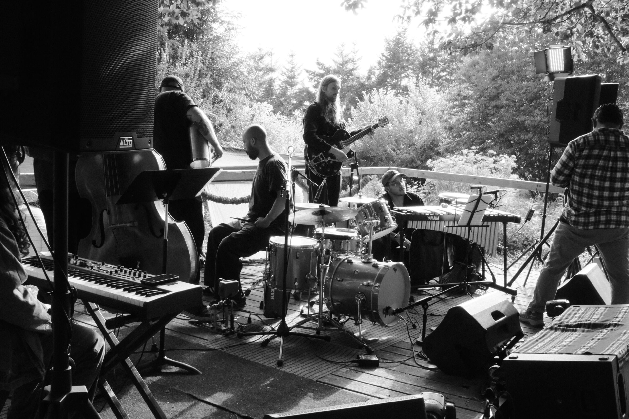 Musicians perform on an outdoor stage with instruments, including drums, keyboard, and guitar, during daytime. People are watching.