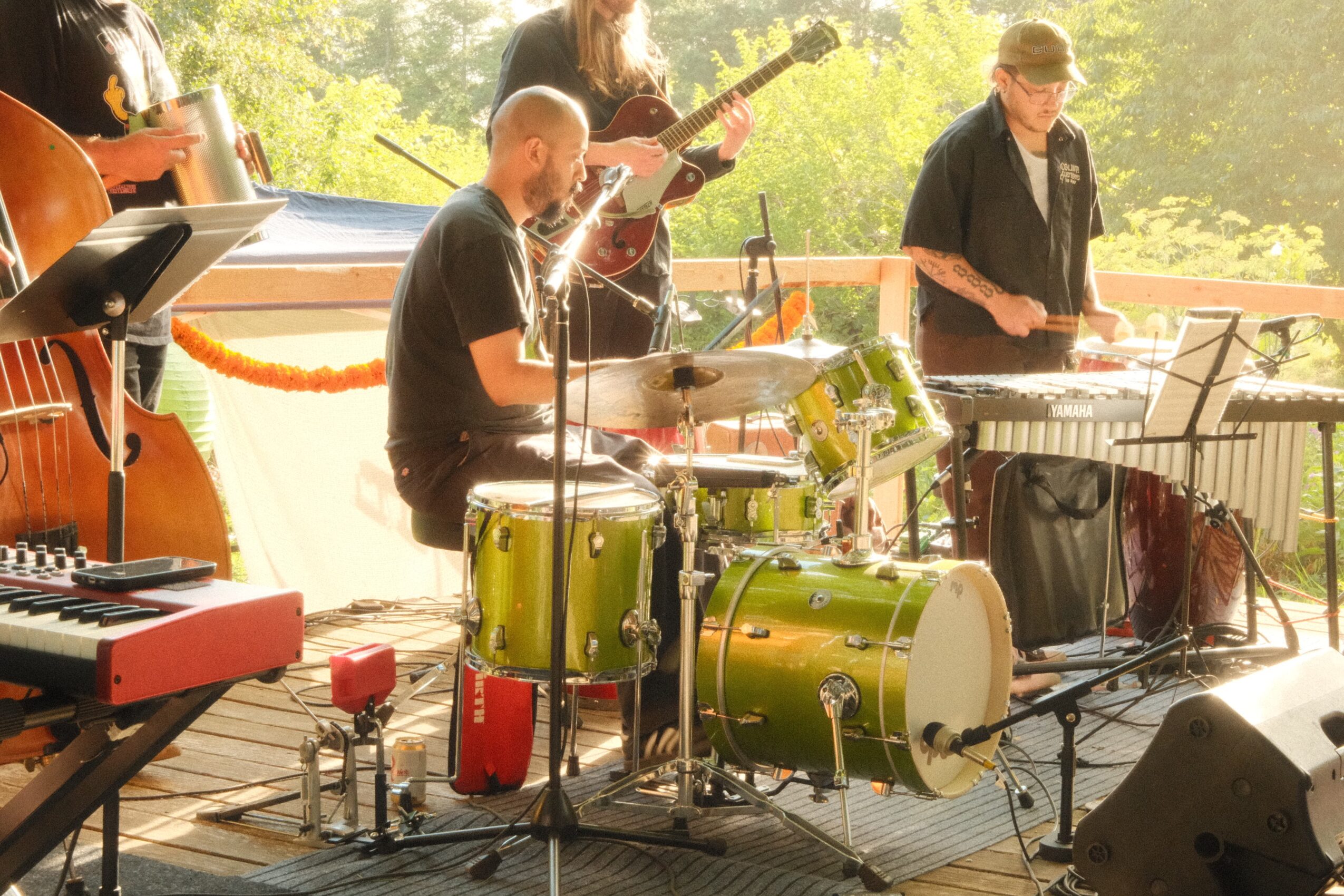 Group of musicians playing instruments outdoors, including drums, keyboard, and guitar, with trees and sunlight in the background.