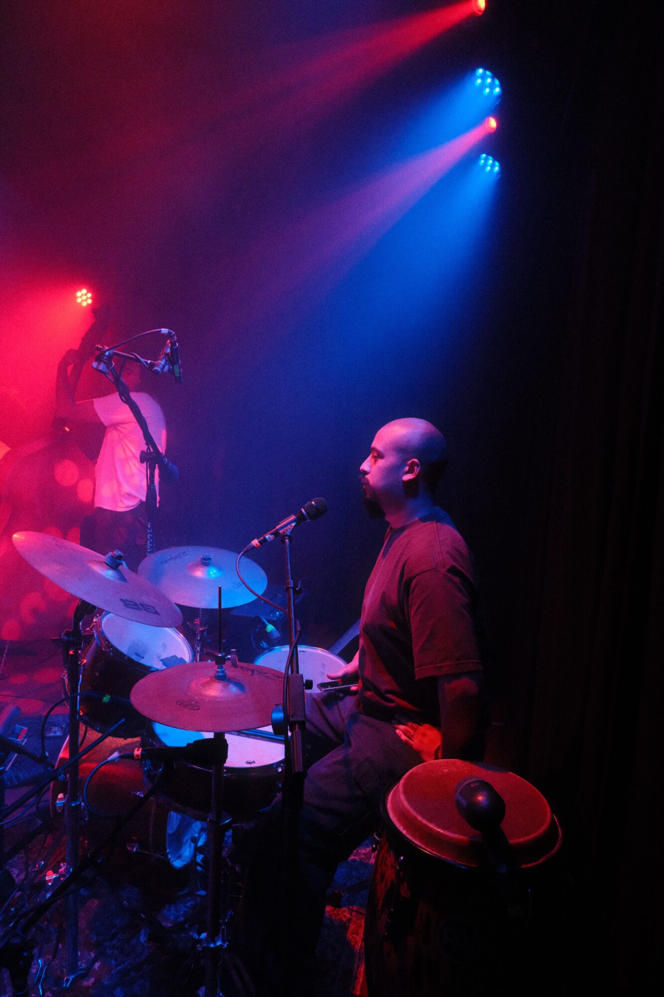 Performer with a hat and glasses on stage with colorful lighting, playing drums or percussion instruments.