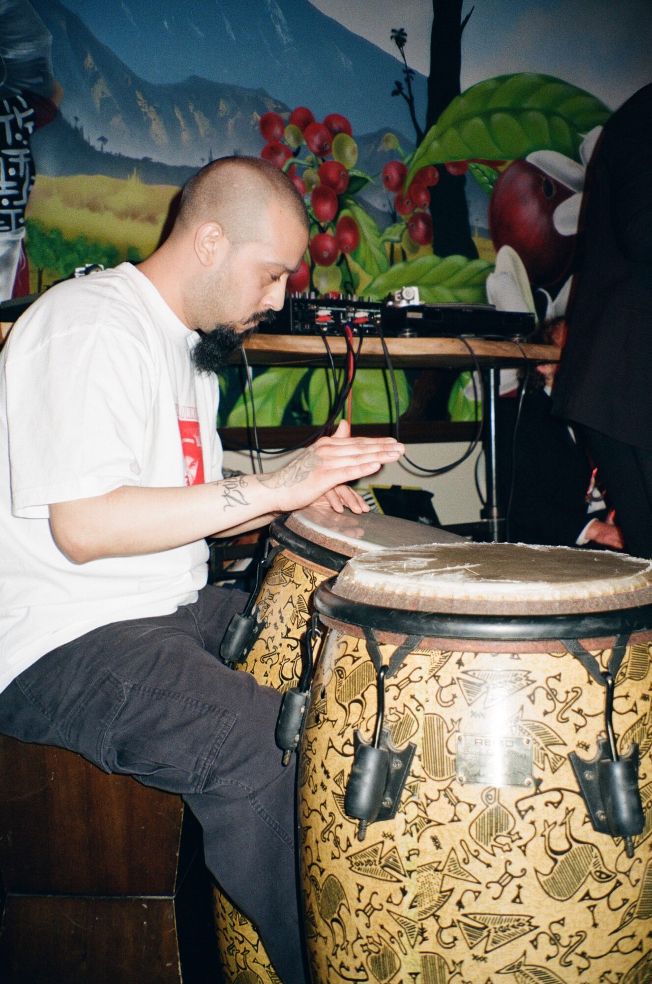 Man playing a large drum with a painted outdoor scene in the background, wearing a white shirt and black pants.